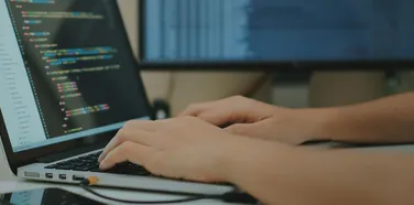 Developer typing on a laptop showing source code, with a second monitor behind it