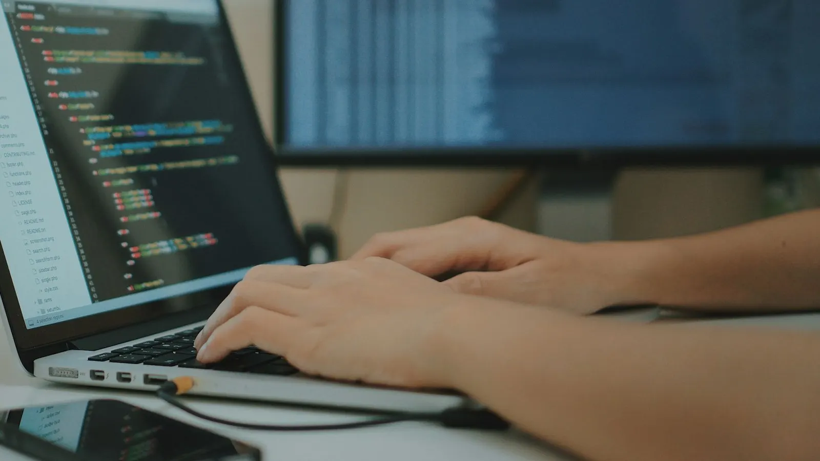 Developer typing on a laptop showing source code, with a second monitor behind it